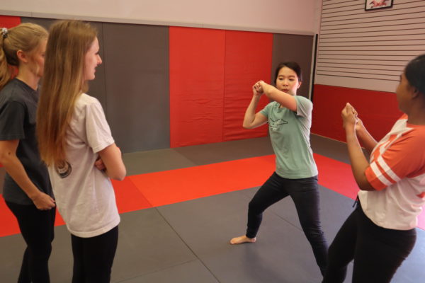 A young woman demonstrates a defensive stance while classmates watch during a self-defense class on red and grey mats